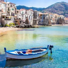 Bateau blanc et bleu ancré dans les eaux cristallines de la plage de Cefalù, avec des maisons colorées en arrière-plan.