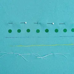 Tissu bleu turquoise avec des épingles, des ronds verts, des traits bleus, jaunes et des fils blancs.