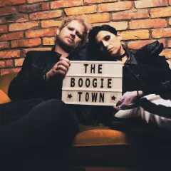 Para siedzi na kanapie, trzymając tabliczkę z napisem "THE BOOGIE TOWN".