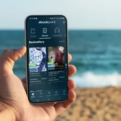 Na smartfonie w dłoni wyświetla się aplikacja ebookpoint z bestsellerami. Idealny poradnik przetrwania na wakacjach, by nie nudzić się na plaży.