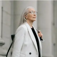 Donna con lunghi capelli grigi, occhiali e un cappotto bianco elegante, con uno sfondo architettonico in tono grigio.