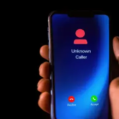 Dłoń trzyma smartfon z ekranem blokady połączeń od nieznanego numeru. Możesz odrzucić lub odebrać. Dowiedz się, jak zastrzec numer.