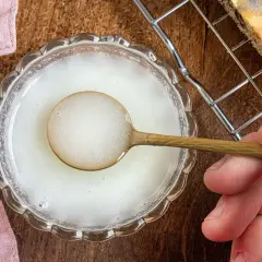 Ein Löffel mit zuckerguss ohne puderzucker wird über eine Schüssel gehalten. Daneben liegt ein Stück Kuchen auf einem Gitter.