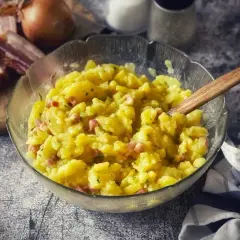 Schüssel mit warmem Kartoffelsalat, garniert mit Speck und Kräutern. Daneben liegen Zwiebeln und Speckstreifen auf einem Holzbrett.