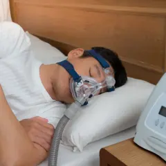 Mann schläft mit CPAP-Maske, um zu wenig Sauerstoff im Schlaf Symptome zu behandeln. Ein Gerät steht daneben.