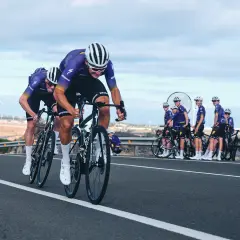 Cykla på Gran Canaria i tävlingsform. Ett cykellag i lila dräkter susar fram på en asfalterad väg med vindkraftverk i bakgrunden.