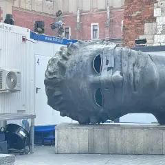 Monumentalna głowa na Rynku w Krakowie, z pustymi oczodołami, stoi obok białych kontenerów.