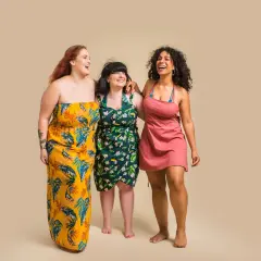 Trzy uśmiechnięte kobiety w kwiecistych sukienkach plus size. Jedna w żółtej, długa, druga w zielonej, krótka, trzecia w różowej, krótka.