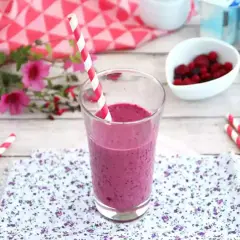Ett glas med en lila indisk yoghurtdryck, dekorerad med ett rött och vitt randigt sugrör. Bredvid finns en skål med bär och yoghurtburkar.