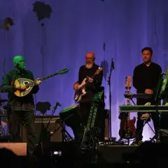 Zespół Dead Can Dance na koncercie. Muzycy grają na instrumentach, w tle fioletowa kurtyna z liśćmi.