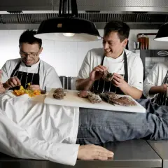 Szefowie kuchni w atelier amaro żartują, jeden z nich leży na blacie, podczas gdy inni przygotowują mięso.