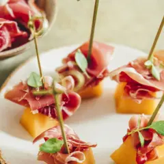 Une présentation originale de melon et jambon, des brochettes savoureuses sur une assiette blanche, avec un melon entier sur le côté.