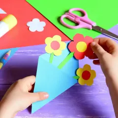 Dziecko tworzy laurkę dla nauczyciela na koniec roku. Kolorowe kwiatki z papieru wkładane są do niebieskiej koperty.