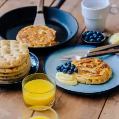 Fattiga riddare med blåbär och vaniljsås på en träyta. En stapel tunna pannkakor och ett glas apelsinjuice.