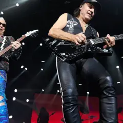 Dwóch gitarzystów na scenie, jeden w skórzanej kurtce i niebieskich spodniach, drugi w czarnej kamizelce i skórzanych spodniach. Ich gra wywołuje "wind of change" tłumaczenie.