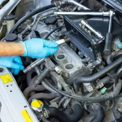 Mano con guante azul sosteniendo una bujía, lista para el cambio de bujías en el motor de un coche. Herramientas a la vista.