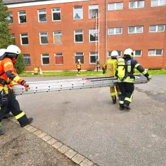 Feuerwehr-Messe: Einsatzkräfte transportieren eine Leiter vor einem Backsteingebäude. Eine Person steigt bereits eine Leiter zum Fenster empor.