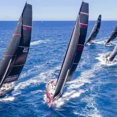 Diverse barche a vela nere con vele rosse e bianche competono in una regata, solcando le onde blu con determinazione.