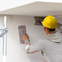 Pracownik w żółtym kasku wykonuje tynkowanie ścian, używając kielni do rozprowadzania masy.