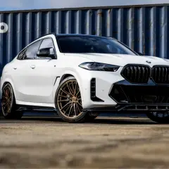 Białe BMW X6 z brązowymi felgami, na których zamontowano opony Imperial, prezentuje się imponująco na tle niebieskiego kontenera.