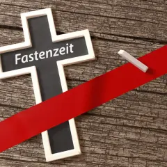 Kreuz mit der Aufschrift "Fastenzeit" auf Holzuntergrund, durchzogen von einem roten Band. Ein Stück Kreide liegt daneben.