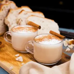 Två koppar krämig chai latte med kanelstänger och strödd kanel. Brödskivor och kanelstänger i bakgrunden.