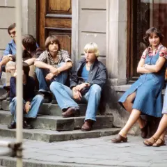 Grupa młodych ludzi w dżinsach siedzi na schodach przed budynkiem.