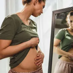 Mujer se mira al espejo, tocándose el abdomen con preocupación por la hinchazón después de comer.