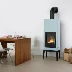 Ein hellblauer Bullerjan B3 Kaminofen mit brennendem Feuer neben einem Holztisch mit Geschirr und Stühlen.