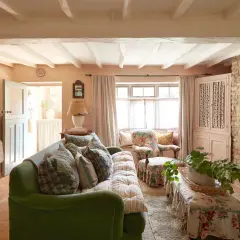 Cozy living room with a green sofa, floral armchair, and charming granny decor. Exposed beams and stone accents add rustic warmth.