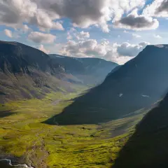 En dramatisk dalgång längs Kungsleden, med solstrålar som bryter igenom molnen och lyser upp den gröna fjällheden.