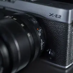 Srebrny aparat Fujifilm X-E2 z obiektywem. Widoczny napis "X-E2" i pokrętło wyboru trybu.