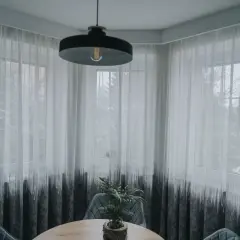 Nowoczesny karnisz na wykusz z białymi zasłonami z czarnym wzorem, tworzący przytulną atmosferę.