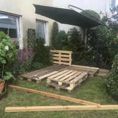 Ogród z meblami z palet. Podłoga z palet w ogrodzie czeka na wykończenie. Obok zielony parasol i roślinność.