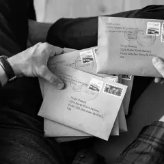 Dłonie trzymają stos brązowych kopert z adresami, znaczkami i pieczątkami pocztowymi. Jedna z kopert jest zaadresowana na "Paige Newell", sugerując odbiór poste restante.