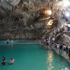 Menschen schwimmen und stehen im türkisfarbenen Wasser einer Höhle in Valladolid, Mexiko. Treppen führen zu einer Plattform, auf der sich weitere Besucher aufhalten.