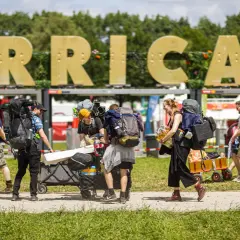 Besucher mit Rucksäcken und Gepäckwagen auf dem Hurricane Zeltplatz. Große goldene Buchstaben "HURRICANE" im Hintergrund.