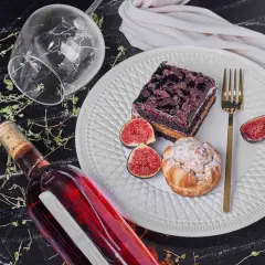 Un bicchiere rovesciato, una bottiglia di vino rosso e un piatto con torta al cioccolato, bignè, fichi e posate dorate, un'invitante composizione per gustare vini da dessert.