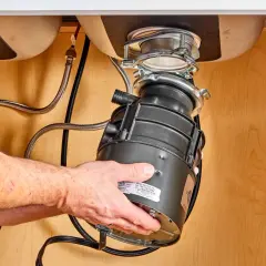 Hands guide a new garbage disposal into place under a sink, part of a garbage disposal installation.