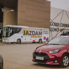 Autobus i czerwony samochód z napisem "JAZDAM SZKOŁA JAZDY" przed budynkiem GOSKIE CENTRUM GOWO-WYSTAWIENNICZE.