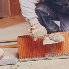 Budowlaniec w rękawiczkach nakłada zaprawę na pustaki, przygotowując je do murowania.