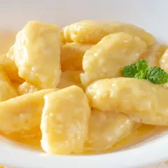 Pyszne, złociste leniwe pierogi w białej miseczce, polane masłem i ozdobione listkiem mięty. Idealne, gdy zastanawiasz się, jak zrobić leniwe pierogi.