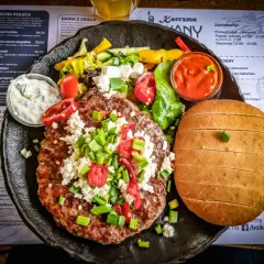 Soczysty kotlet z dodatkami i bułką, serwowany z sosem. Prawdziwa uczta kuchni bałkańskiej.