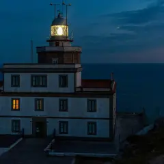 Faro de Finisterre iluminado al anochecer, con el mar oscuro de fondo. Una luz cálida brilla en una ventana.