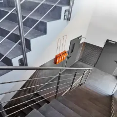 Moderner Treppenaufgang mit Metallgeländern und grauen Stufen. An der Wand hängen orangefarbene Geräte, die Teil des Rauchabzug Treppenhaus Systems sind.