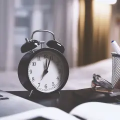 Budzik, laptop, okulary i przybory piśmiennicze na biurku.