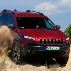 Czerwony Jeep Cherokee Trailhawk z impetem przemierza piaszczysty teren, wzbijając tumany piasku.