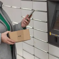 Kobieta z paczką w ręku korzysta z telefonu przy automacie InPost. Szybkie przekierowanie paczki jest teraz łatwiejsze.