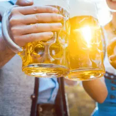Zwei Hände halten je ein **Mass Bier** und stoßen an. Eine Brezel liegt in der anderen Hand.