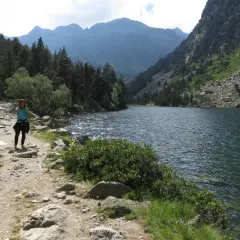 Una mujer camina por un sendero rocoso junto a un estany llong, rodeado de montañas y pinos.